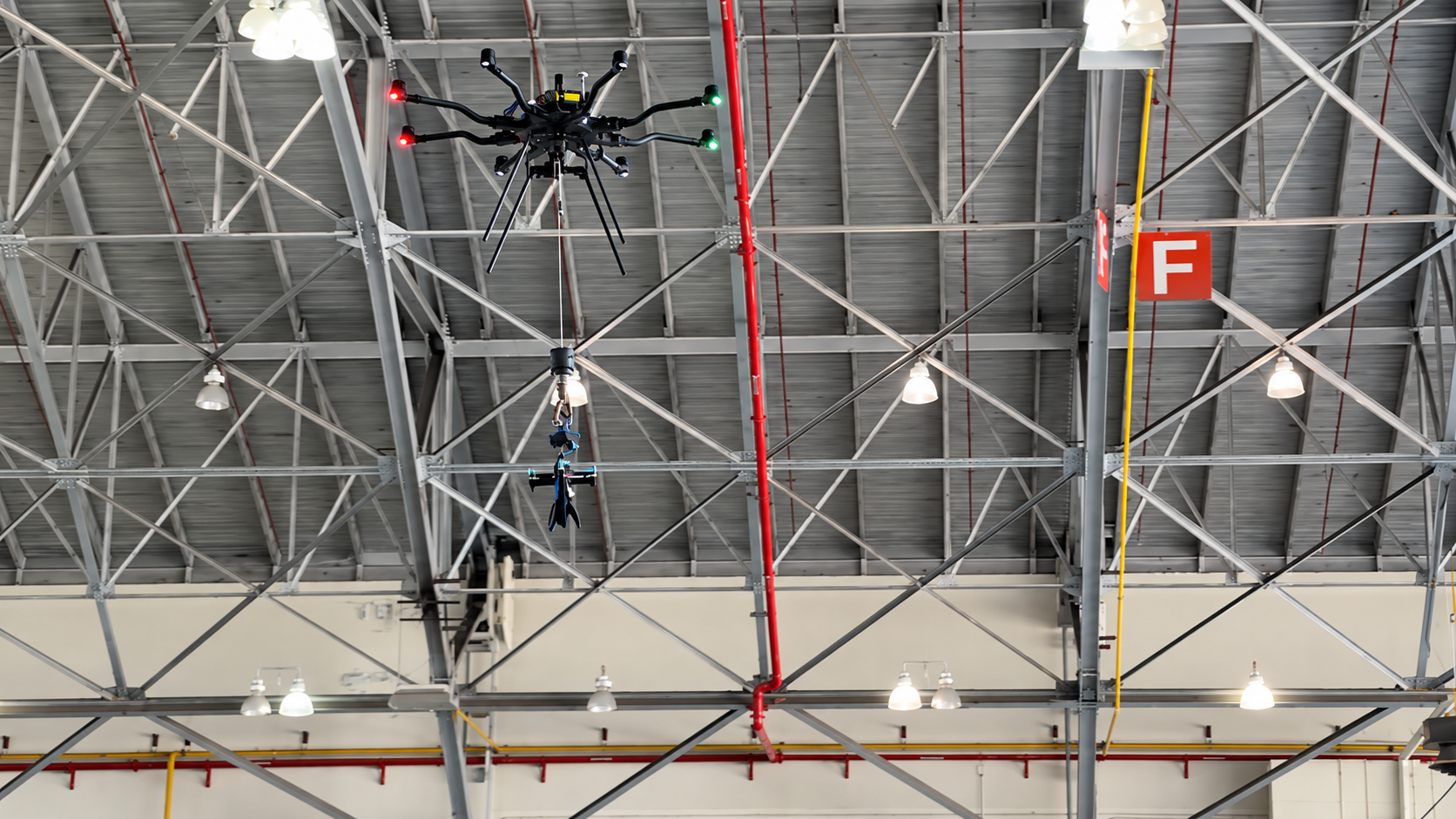 Carrier multirotor holding the bullet drone beneath its release rig during indoor drop-test preparation